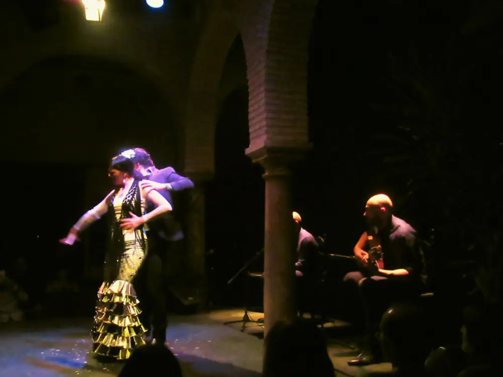 Seville's Bienal de Flamenco 2026 2 Flamenco show showing female dancer in golden dress with guitarists in the background in the shadows - andalucia.org - © TURISMO ANDALUZ
