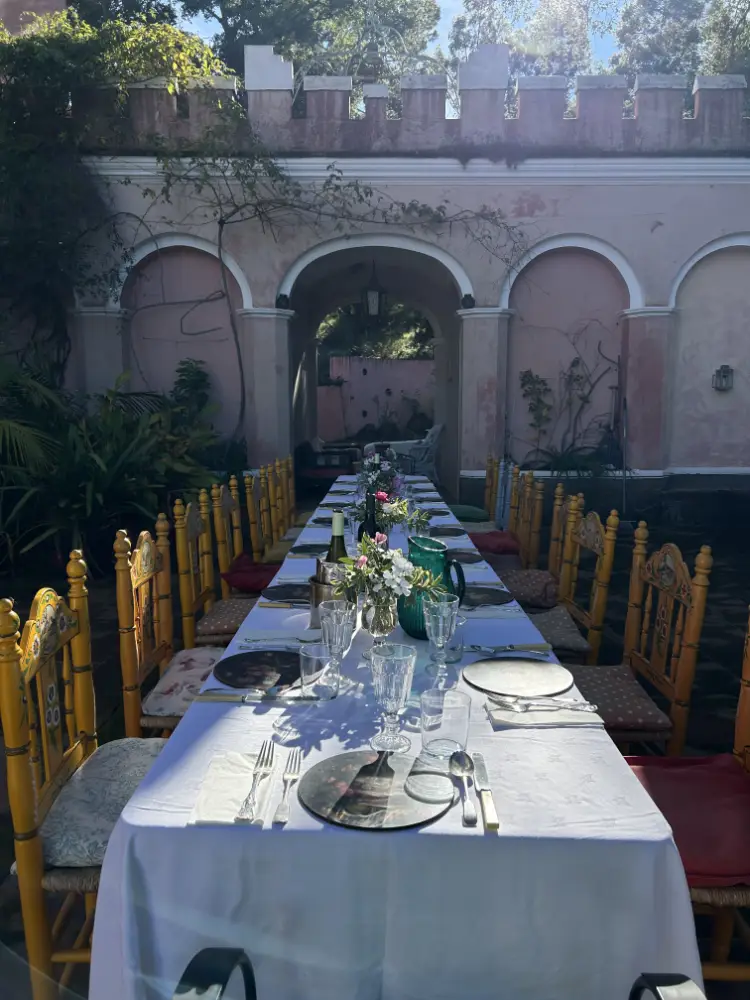 Andalucia Writers Retreat - Long outdoor dining table elegantly set with white linens, glassware, floral arrangements and yellow chairs in a sunlit courtyard.