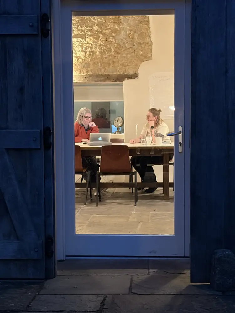 Dorset Writers Retreat - Two women writing together at a table inside a warmly lit rustic room, viewed through a glass-panelled doorway