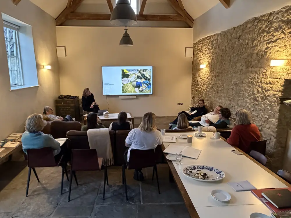 Writers retreat participants listening to a presentation in a rustic meeting room with exposed stone walls, wooden beams and a large shared table.