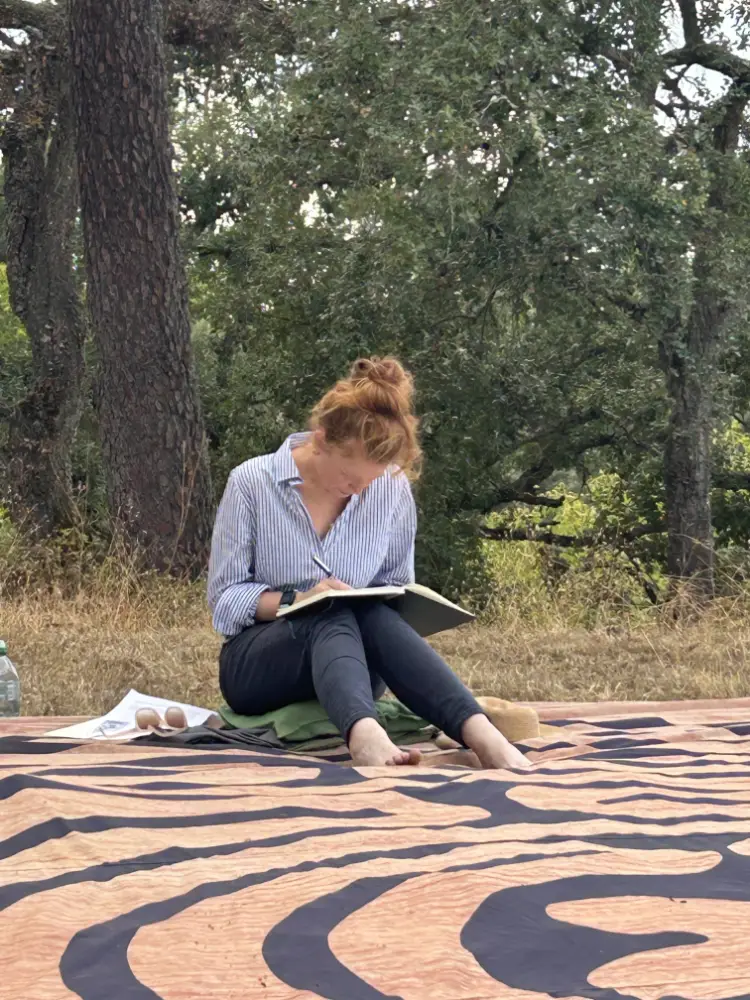 A women sitting barefoot on a patterned outdoor mat, writing in a notebook beneath trees in a woodland setting